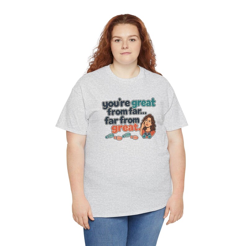 May include: A light gray short-sleeve t-shirt featuring a graphic print with the text "you're great from far... far from great." The design includes a cartoon illustration of a person looking through binoculars and several small footprint icons.