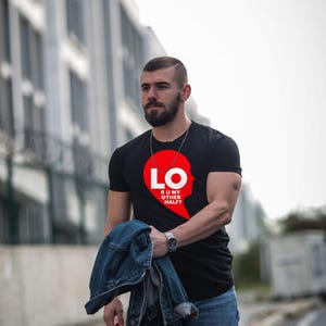 Puede incluir: Una camiseta negra de manga corta con un gr&aacute;fico rojo en forma de coraz&oacute;n en el pecho. El gr&aacute;fico contiene el texto blanco LO y el texto blanco m&aacute;s peque&ntilde;o R U MY OTHER HALF? dispuestos verticalmente en el coraz&oacute;n.