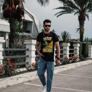 May include: A black short-sleeved t-shirt featuring a graphic of a person rooted as a tree with the text I AM STUCK AS A TREE WHILE THEY GET TRILLIONS, paired with blue denim jeans and white sneakers.