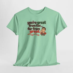 May include: A light green short-sleeve t-shirt featuring a graphic of a person looking through binoculars with the text "you're great from far... far from great." and small red footprints printed below the text on the front.