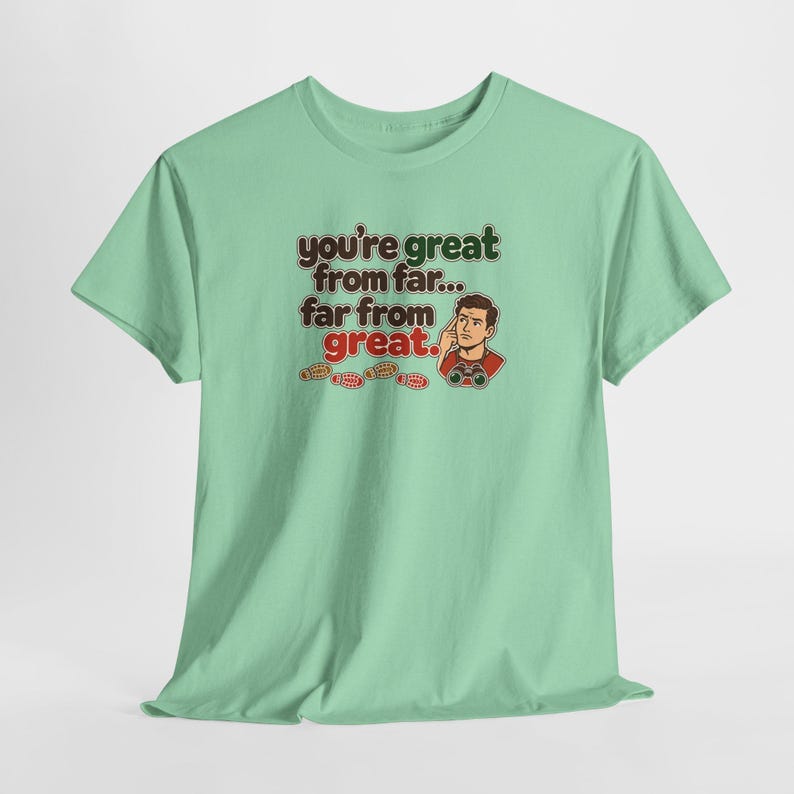 May include: A light green short-sleeve t-shirt featuring a graphic of a person looking through binoculars with the text "you're great from far... far from great." and small red footprints printed below the text on the front.