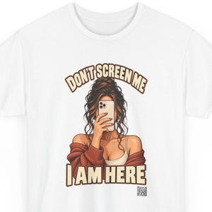 K&ouml;nnte beinhalten: Ein wei&szlig;es T-Shirt mit einer grafischen Illustration einer Person mit braunen Haaren in einem unordentlichen Dutt, die eine rostfarbene Strickjacke &uuml;ber einem wei&szlig;en Tanktop tr&auml;gt und ein Smartphone vor das Gesicht h&auml;lt.