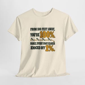 May include: A cream-colored short-sleeve t-shirt featuring black and gold text that reads: FROM 100 FEET AWAY, YOU'RE 100%. SADLY, EVERY FOOT CLOSER KNOCKS OFF 1%. The design includes a horizontal line with small footprint icons.