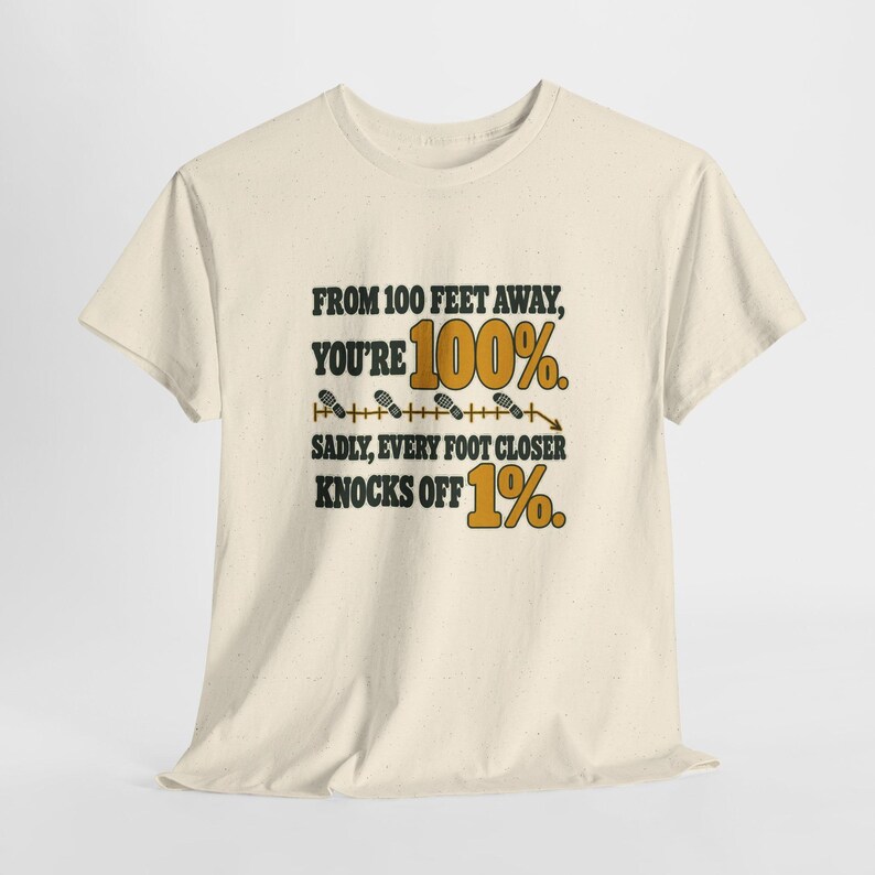 May include: A cream-colored short-sleeve t-shirt featuring black and gold text that reads: FROM 100 FEET AWAY, YOU'RE 100%. SADLY, EVERY FOOT CLOSER KNOCKS OFF 1%. The design includes a horizontal line with small footprint icons.