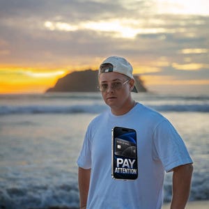 May include: A white t-shirt featuring a graphic print of a smartphone displaying a credit card and the text PAY ATTENTION. The shirt is worn with a light-colored baseball cap, wire-rimmed glasses, and a watch on the wrist.