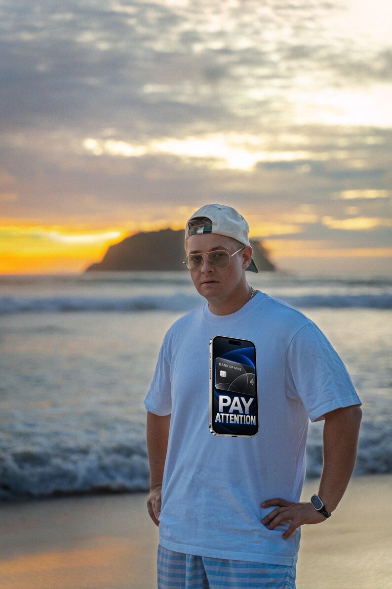 May include: A white t-shirt featuring a graphic print of a smartphone displaying a credit card and the text PAY ATTENTION. The shirt is worn with a light-colored baseball cap, wire-rimmed glasses, and a watch on the wrist.