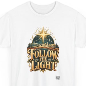 May include: A white t-shirt featuring a graphic print of a golden star shining over a landscape with a small town. Below the illustration, the text reads "FOLLOW THE LIGHT" in a decorative gold font. A small QR code is visible in the bottom right.