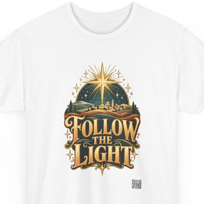 May include: A white t-shirt featuring a graphic print of a golden star shining over a landscape with a small town. Below the illustration, the text reads "FOLLOW THE LIGHT" in a decorative gold font. A small QR code is visible in the bottom right.