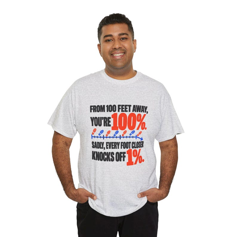 May include: A light gray short-sleeve t-shirt featuring black and red text that reads: FROM 100 FEET AWAY, YOU'RE 100%. SADLY, EVERY FOOT CLOSER KNOCKS OFF 1%. A blue line with alternating red and blue footprint icons is included.