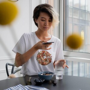 Puede incluir: Una camiseta blanca de manga corta con un gr&aacute;fico circular de un drag&oacute;n estilizado en tonos rojos, naranjas y dorados. El dise&ntilde;o est&aacute; centrado en el pecho de la prenda, confeccionada en tela blanca lisa con cuello redondo.