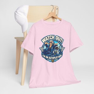 May include: A light pink t-shirt featuring a graphic of a pilot and a robot in a cockpit with the text WATCH OUT! I'M ON AUTOPILOT NOW. The shirt is displayed on a wooden hanger with a white baseball cap hanging on the back of the chair.