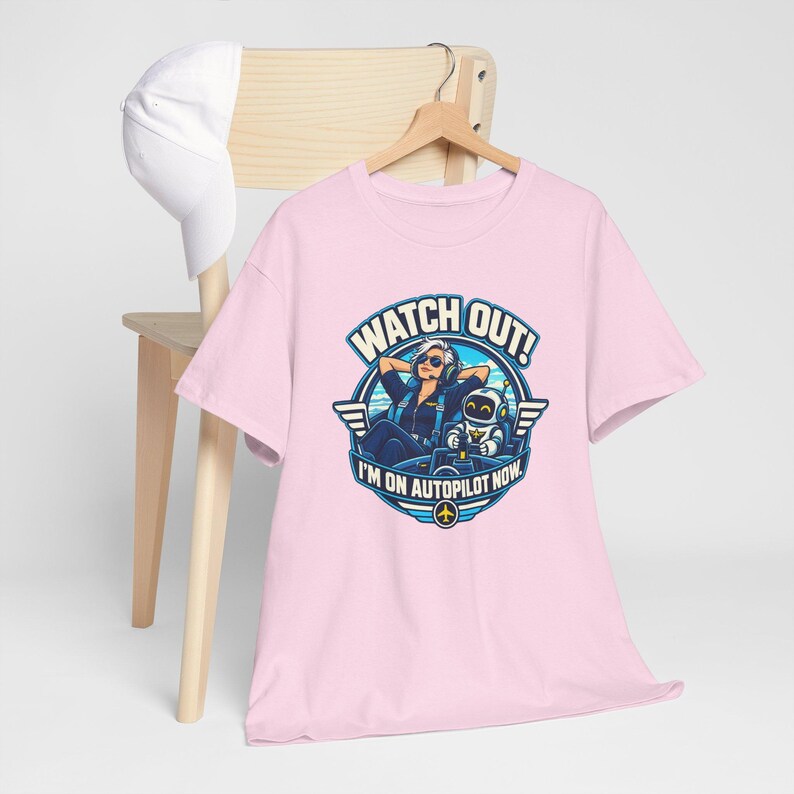 May include: A light pink t-shirt featuring a graphic of a pilot and a robot in a cockpit with the text WATCH OUT! I'M ON AUTOPILOT NOW. The shirt is displayed on a wooden hanger with a white baseball cap hanging on the back of the chair.