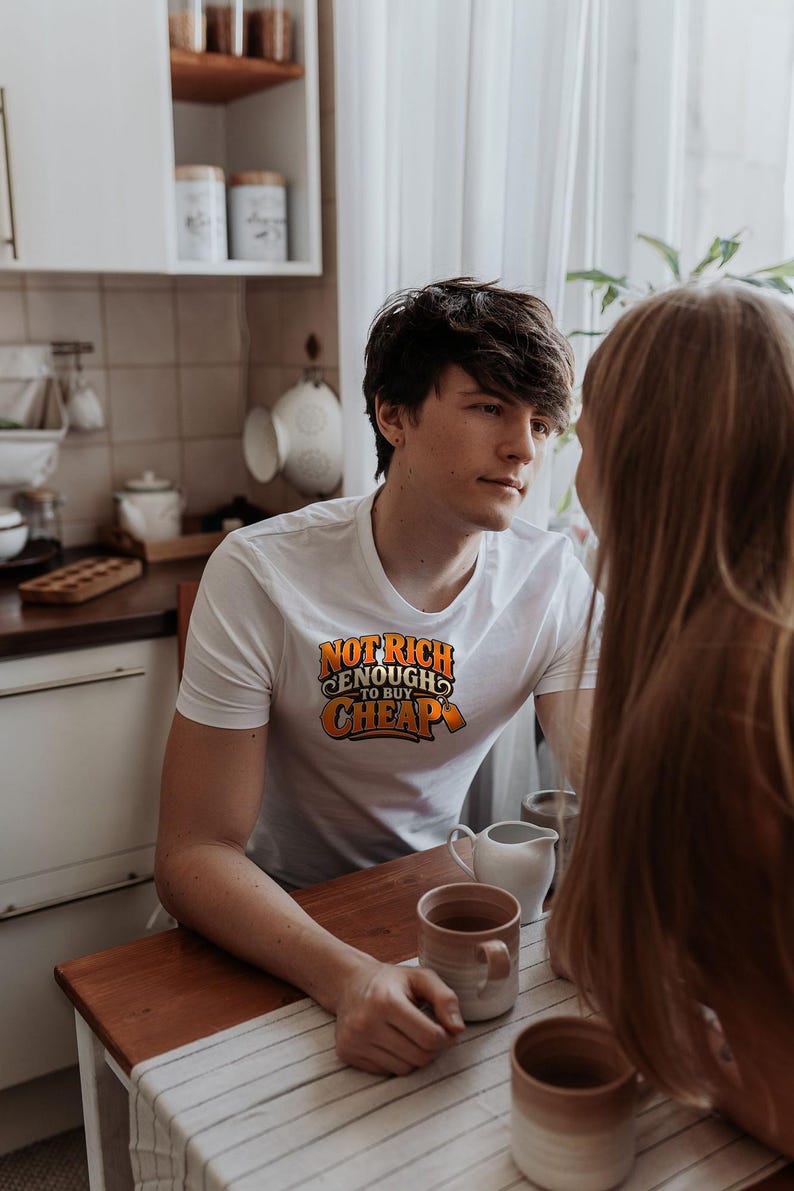 May include: A white short-sleeved t-shirt featuring the orange and black text "NOT RICH ENOUGH TO BUY CHEAP" printed on the chest. The shirt has a crew neckline and is made of a smooth fabric, shown in a kitchen setting with mugs on a table.
