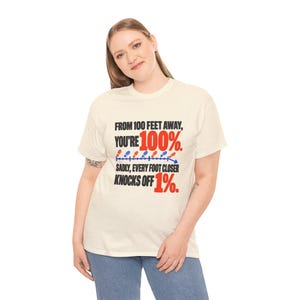 May include: A cream-colored short-sleeve t-shirt featuring black and red text that reads: FROM 100 FEET AWAY, YOU'RE 100%. SADLY, EVERY FOOT CLOSER KNOCKS OFF 1%. A graphic of small red and blue footprints follows a downward arrow.