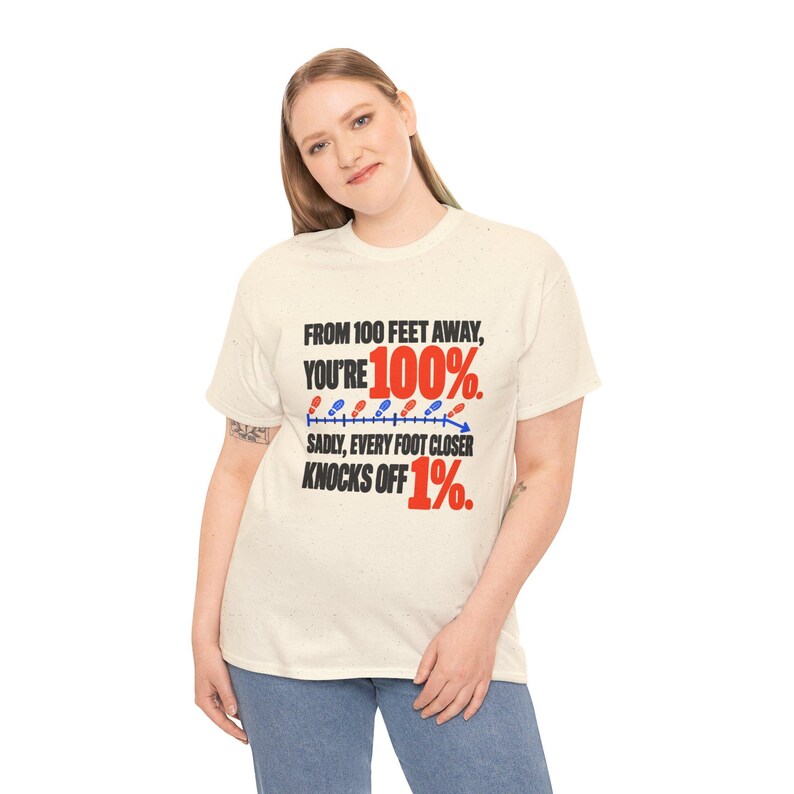 May include: A cream-colored short-sleeve t-shirt featuring black and red text that reads: FROM 100 FEET AWAY, YOU'RE 100%. SADLY, EVERY FOOT CLOSER KNOCKS OFF 1%. A graphic of small red and blue footprints follows a downward arrow.