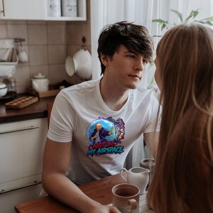 Może przedstawiać: Biały t-shirt z kr&oacute;tkim rękawem z grafiką przedstawiającą łysą postać w niebieskiej bańce z fioletowym dymem oraz napisem NOT IN MY AIRSPACE niebiesko-czerwonymi literami umieszczonym na klatce piersiowej.