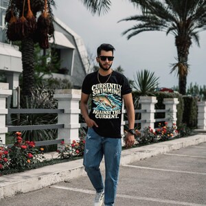 May include: A black short-sleeved t-shirt featuring a graphic of a fish jumping out of blue water with the text "CURRENTLY SWIMMING AGAINST THE CURRENT." The shirt is paired with blue denim jeans and white athletic sneakers.