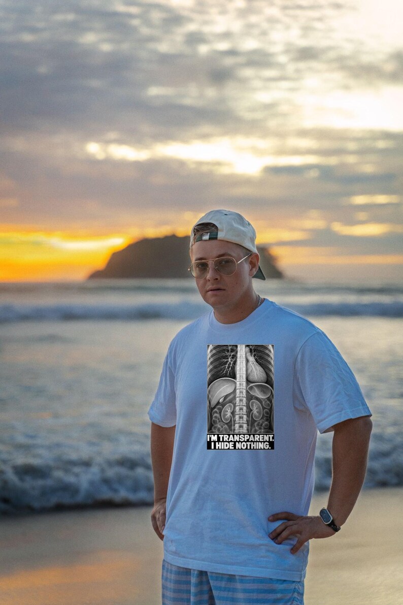 Puede incluir: Una camiseta blanca de manga corta con un gr&aacute;fico en blanco y negro de una radiograf&iacute;a del torso humano. El gr&aacute;fico incluye el texto "I'M TRANSPARENT. I HIDE NOTHING." impreso en una fuente negrita sin remates en la parte inferior.