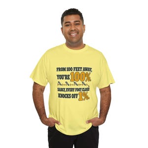 May include: A light yellow short-sleeve t-shirt featuring black and gold text that reads: FROM 100 FEET AWAY, YOU'RE 100%. SADLY, EVERY FOOT CLOSER KNOCKS OFF 1%. The design includes a horizontal line with small footprint icons.