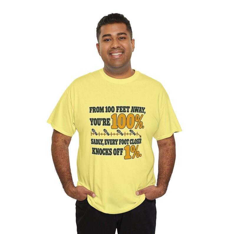 May include: A light yellow short-sleeve t-shirt featuring black and gold text that reads: FROM 100 FEET AWAY, YOU'RE 100%. SADLY, EVERY FOOT CLOSER KNOCKS OFF 1%. The design includes a horizontal line with small footprint icons.