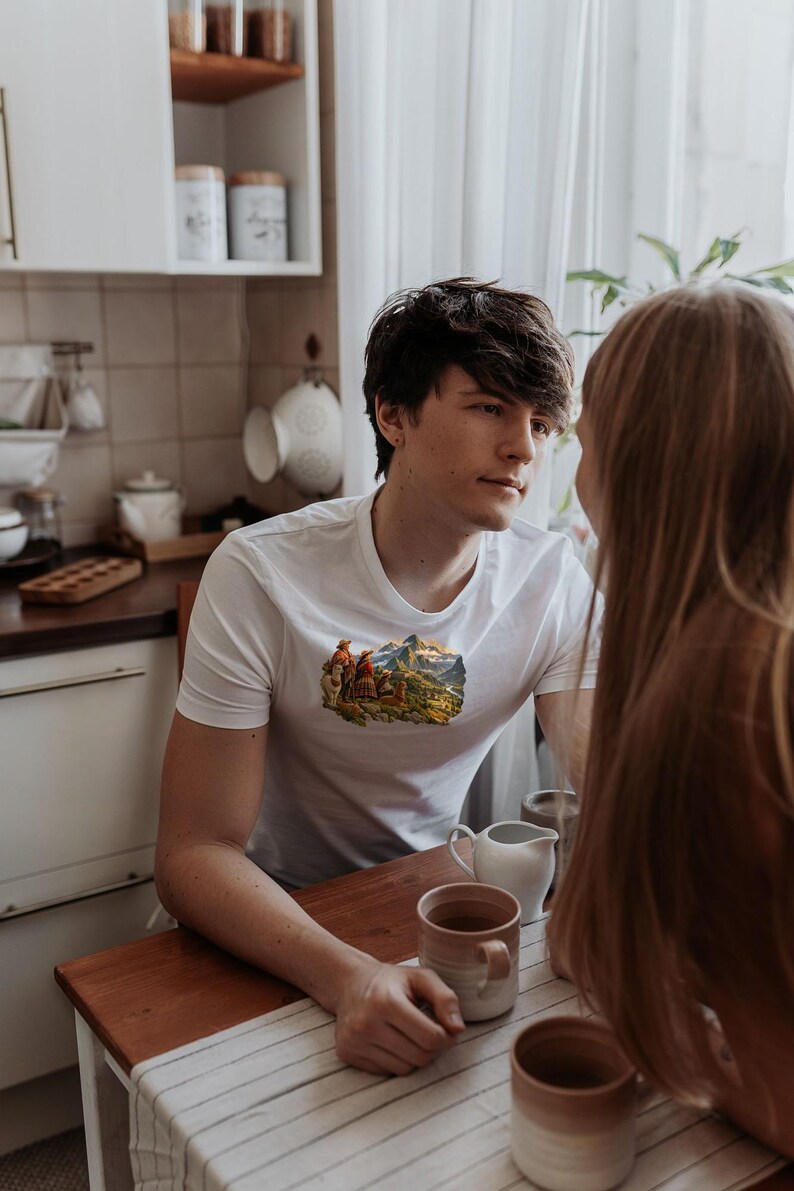 May include: A white short-sleeved t-shirt featuring a small, detailed graphic print on the chest. The graphic depicts a landscape scene with mountains, figures, and animals. The shirt has a classic crew neckline and a fitted cut.