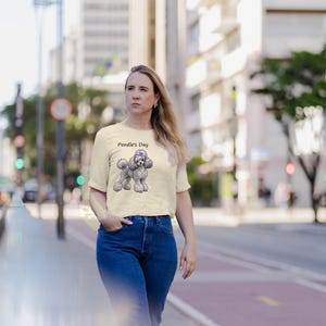 May include: A cream-colored short-sleeve t-shirt featuring a graphic illustration of a grey poodle wearing a purple bow on its head, with the text Poodle's Day printed in black above the dog illustration on the front of the garment.