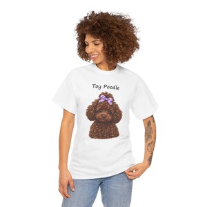 May include: A white short-sleeved t-shirt featuring a graphic of a brown curly-haired toy poodle wearing a light purple bow on its head. The text Toy Poodle is printed in black above the dog illustration on the front of the garment.