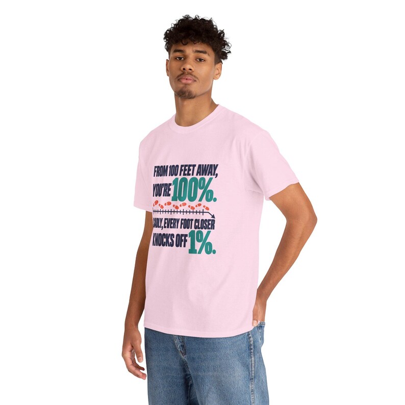 May include: A light pink short-sleeve t-shirt featuring dark blue and teal text that reads: FROM 100 FEET AWAY, YOU'RE 100%. SADLY, EVERY FOOT CLOSER KNOCKS OFF 1%. The design includes a horizontal line with small red footprints above it.