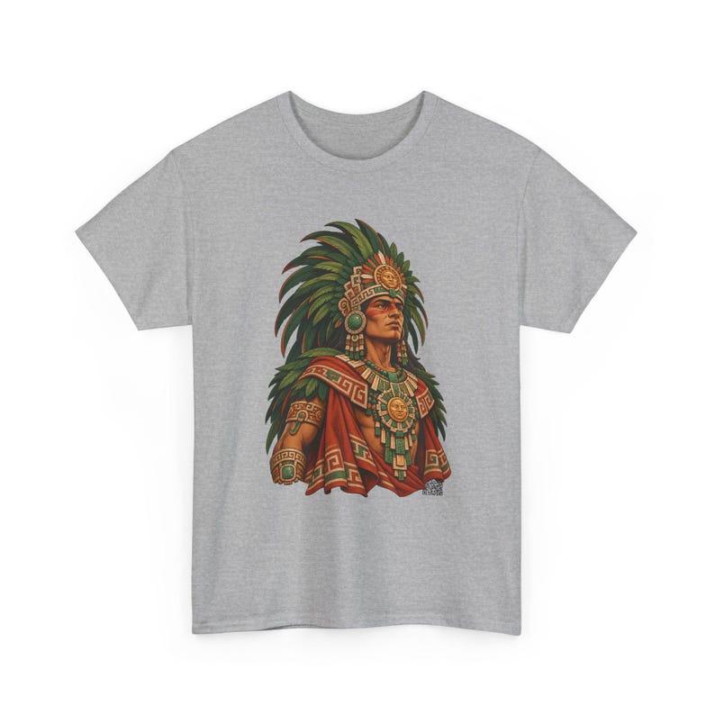 May include: A heather gray t-shirt featuring a graphic print of a figure wearing an elaborate green feathered headdress, a red cape, and gold jewelry with sun motifs. The design includes intricate geometric patterns in green, red, and gold tones.