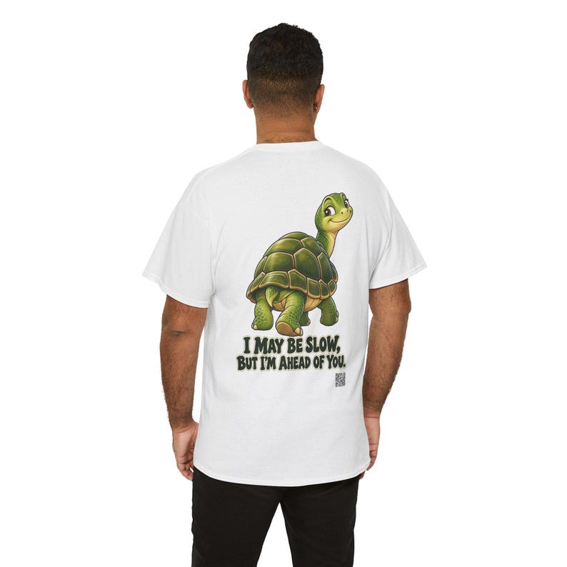 May include: A white short-sleeved t-shirt featuring a graphic of a green turtle looking back over its shoulder. Below the turtle, the text reads, I MAY BE SLOW, BUT I'M AHEAD OF YOU. The design is centered on the back of the garment.