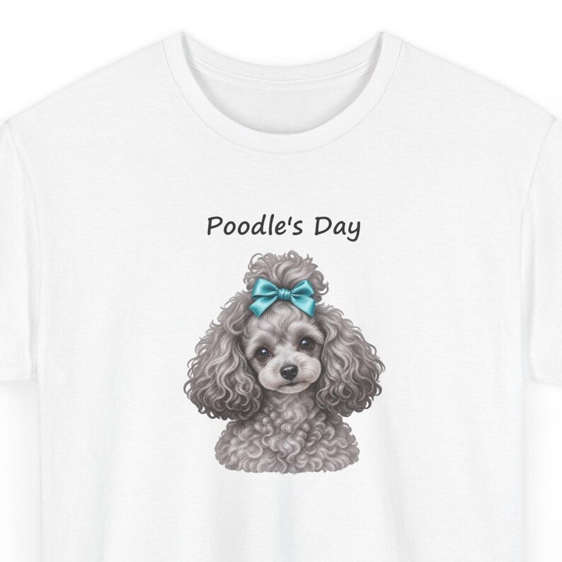 Puede incluir: Una camiseta blanca con el gr&aacute;fico de un caniche gris de pelo rizado con un lazo azul brillante en la cabeza. Sobre el caniche, el texto Poodle's Day est&aacute; impreso en una fuente negra sencilla. El dise&ntilde;o est&aacute; centrado en el pecho.