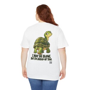 May include: A white t-shirt featuring a graphic of a green turtle looking back over its shoulder. Below the turtle, the text reads, "I MAY BE SLOW, BUT I'M AHEAD OF YOU." A small QR code is printed beneath the text on the back of the garment.