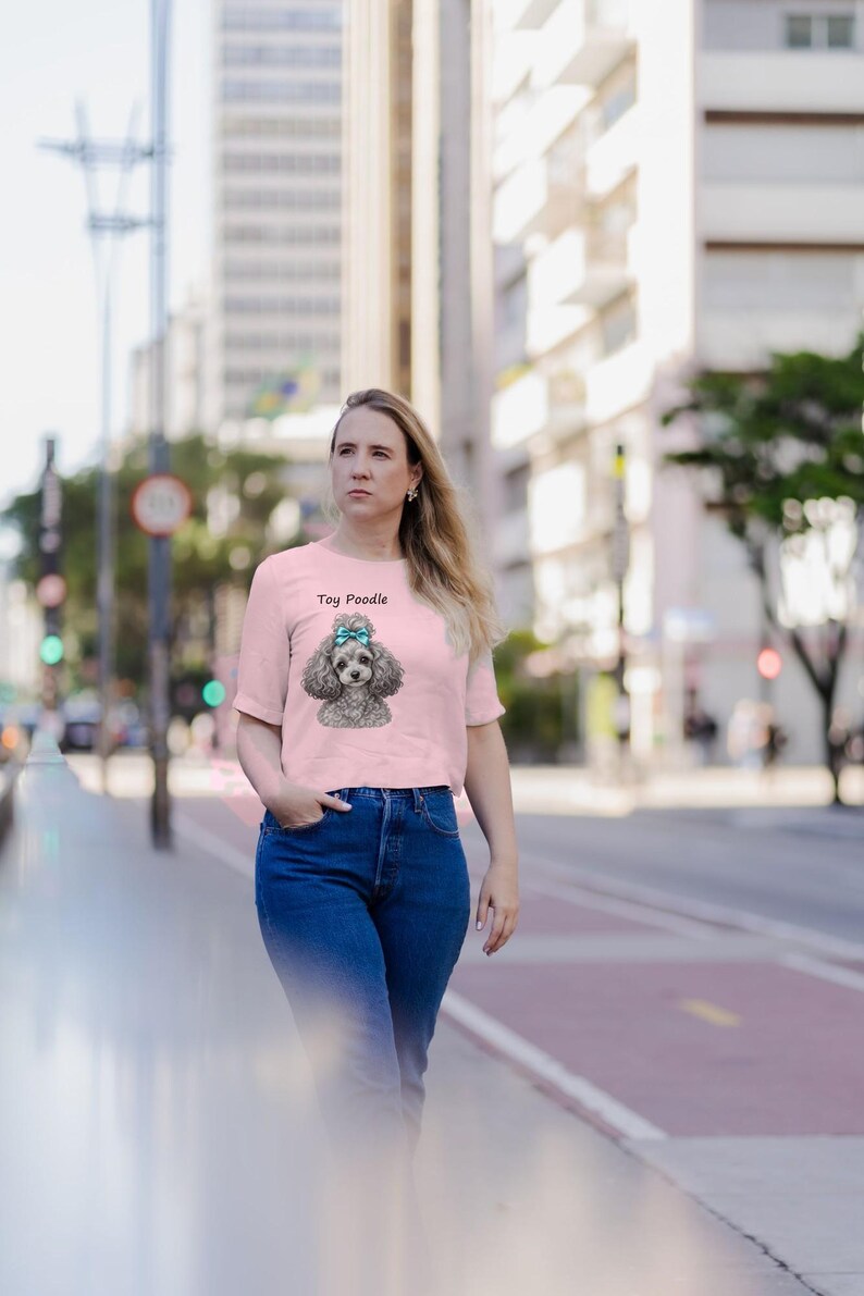 K&ouml;nnte beinhalten: Ein hellrosa T-Shirt mit kurzen &Auml;rmeln und einer grafischen Illustration eines grauen Toypudels mit einer t&uuml;rkisfarbenen Schleife auf dem Kopf. Der Text "Toy Poodle" ist in Schwarz &uuml;ber der Hundeillustration auf dem Kleidungsst&uuml;ck gedruckt.