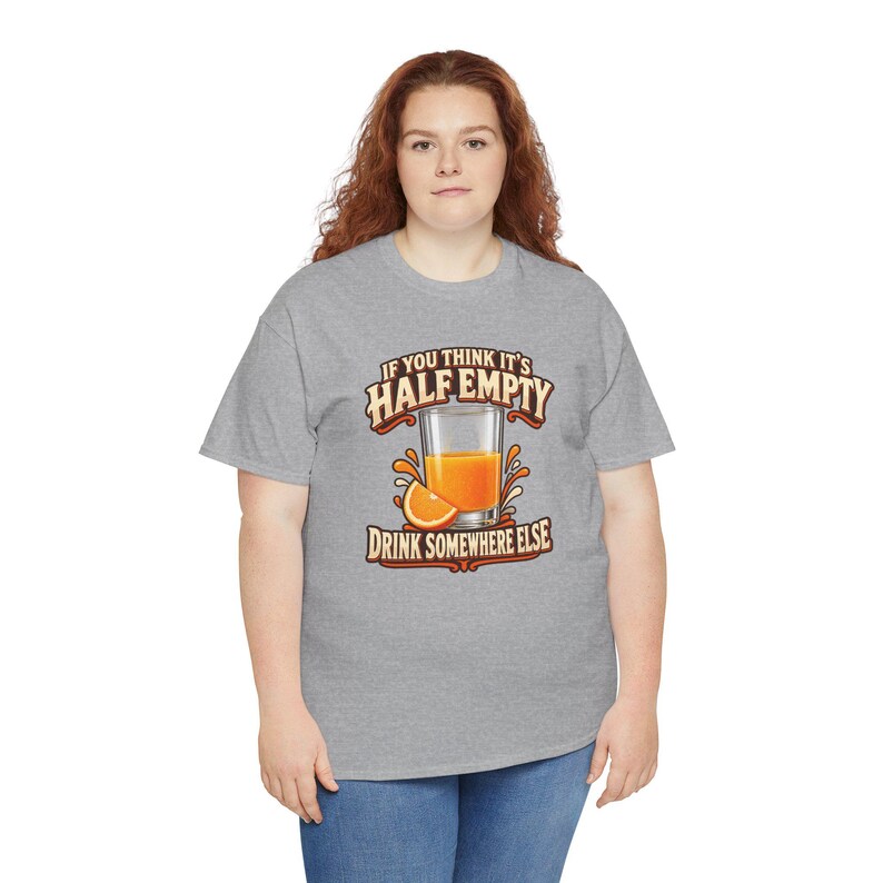 May include: A heather gray t-shirt featuring a graphic of a glass of orange juice with an orange slice. The text reads: IF YOU THINK IT'S HALF EMPTY DRINK SOMEWHERE ELSE. The design uses orange, yellow, and brown tones on the front of the shirt.