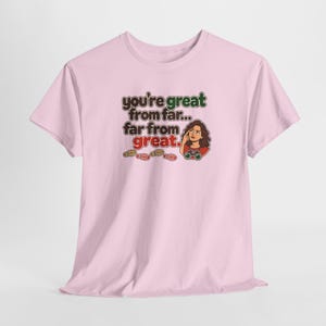 May include: A light pink short-sleeve t-shirt featuring the text "you're great from far... far from great." in brown, green, and red. The design includes a cartoon illustration of a person with binoculars and four small footprints.