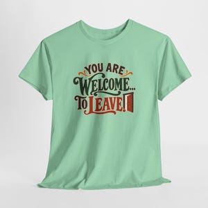 May include: A light mint green short-sleeve t-shirt featuring the dark brown and red text "YOU ARE WELCOME... TO LEAVE!" with a small graphic of an open door next to the word LEAVE, centered on the chest of the garment.