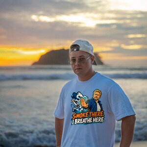 May include: A white short-sleeve t-shirt featuring a graphic illustration of a person with a hand raised in a stop gesture, a cigarette, and blue smoke. The text reads SMOKE THERE. I BREATHE HERE. The design uses blue, orange, and white colors.