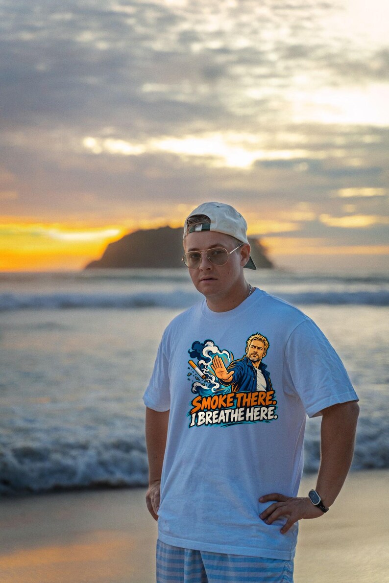 May include: A white short-sleeve t-shirt featuring a graphic illustration of a person with a hand raised in a stop gesture, a cigarette, and blue smoke. The text reads SMOKE THERE. I BREATHE HERE. The design uses blue, orange, and white colors.