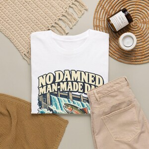 Puede incluir: Una camiseta blanca con un gr&aacute;fico de una presa con agua fluyendo y una trucha en primer plano. El texto "NO DAMNED MAN-MADE DAM" est&aacute; impreso en letras grandes, negritas y de color crema sobre la ilustraci&oacute;n de la presa.