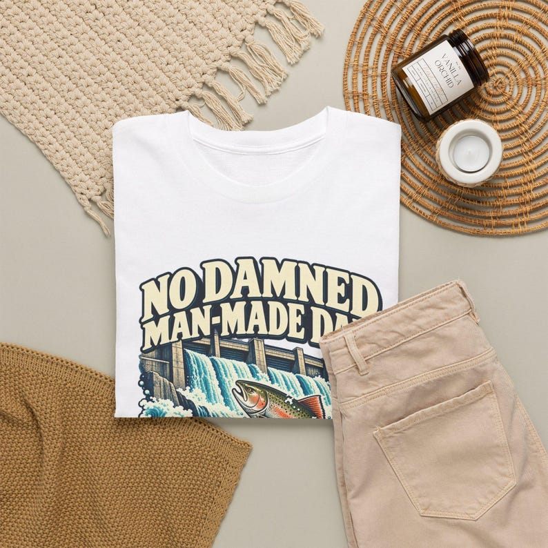 Puede incluir: Una camiseta blanca con un gr&aacute;fico de una presa con agua fluyendo y una trucha en primer plano. El texto "NO DAMNED MAN-MADE DAM" est&aacute; impreso en letras grandes, negritas y de color crema sobre la ilustraci&oacute;n de la presa.