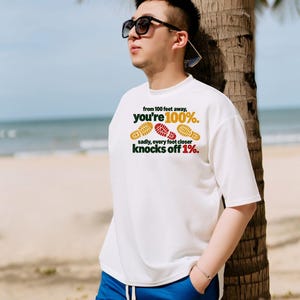 May include: A white short-sleeved t-shirt featuring the text 'from 100 feet away, you're 100%. sadly, every foot closer knocks off 1%.' along with three colorful shoe print graphics in yellow and red.