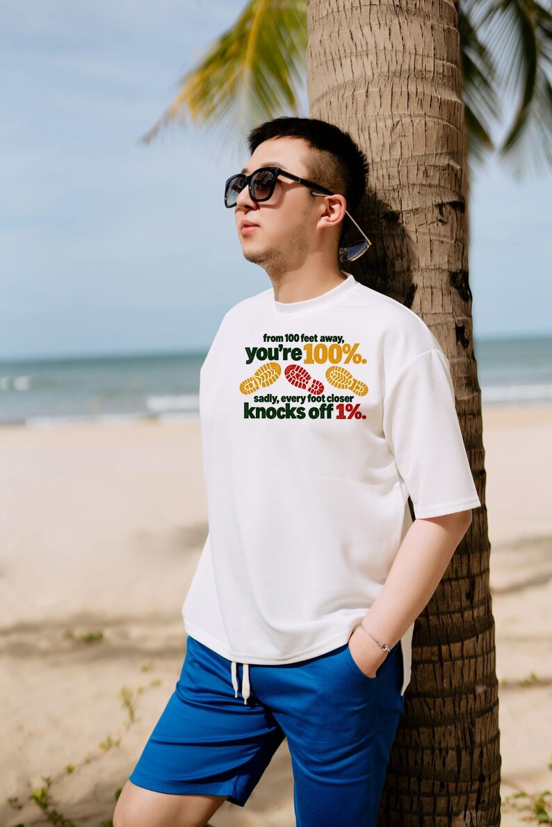May include: A white short-sleeved t-shirt featuring the text 'from 100 feet away, you're 100%. sadly, every foot closer knocks off 1%.' along with three colorful shoe print graphics in yellow and red.