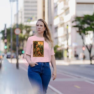 May include: A light pink short-sleeved t-shirt featuring a rectangular graphic print of a person wearing ornate green and gold traditional attire with a feathered headdress, set against a backdrop of a sun and ancient stone structures.