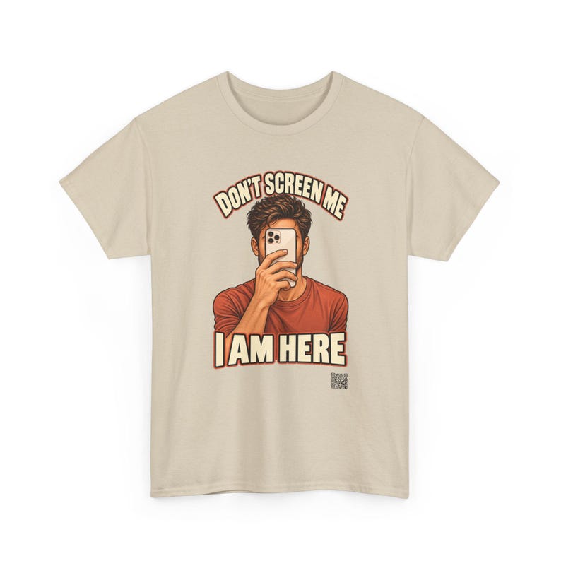 Op de afbeelding: Een beige t-shirt met een afbeelding van een persoon die een smartphone voor het gezicht houdt. De tekst erboven luidt DON'T SCREEN ME en de tekst eronder luidt I AM HERE in vetgedrukte letters. Een kleine QR-code staat rechtsonder gedrukt.