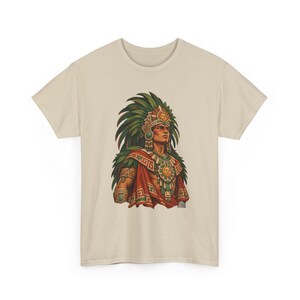 May include: A beige short-sleeve t-shirt featuring a graphic print of a figure wearing a large green feathered headdress, a red cape, and gold sun-themed jewelry. The illustration displays intricate patterns and vibrant colors centered on the chest.