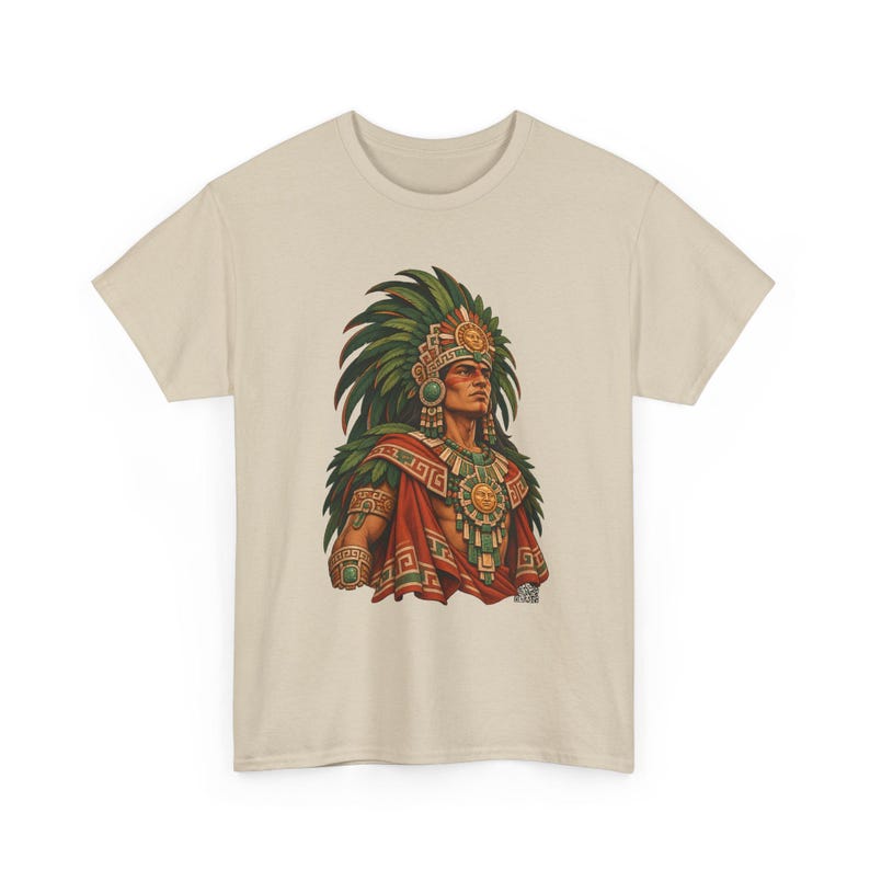 May include: A beige short-sleeve t-shirt featuring a graphic print of a figure wearing a large green feathered headdress, a red cape, and gold sun-themed jewelry. The illustration displays intricate patterns and vibrant colors centered on the chest.