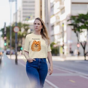 May include: A cream-colored short-sleeve t-shirt featuring a graphic illustration of a brown Pomeranian dog centered on the chest with the word Pomeranian printed in black text above the dog's head, paired with blue denim jeans.