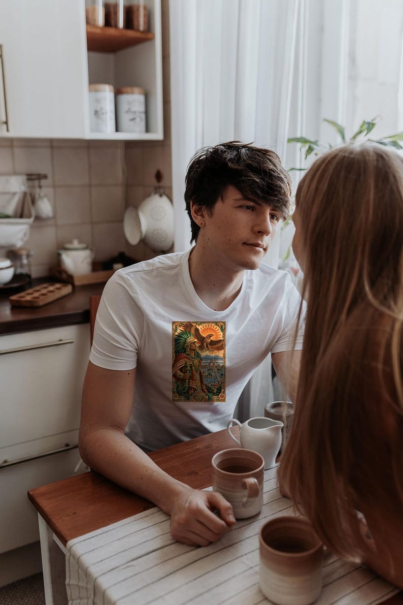 Pu&ograve; includere: Una t-shirt bianca a maniche corte con una stampa grafica rettangolare sul petto. L'illustrazione raffigura una figura in abiti tradizionali decorati con un grande copricapo di piume davanti a un paesaggio urbano.