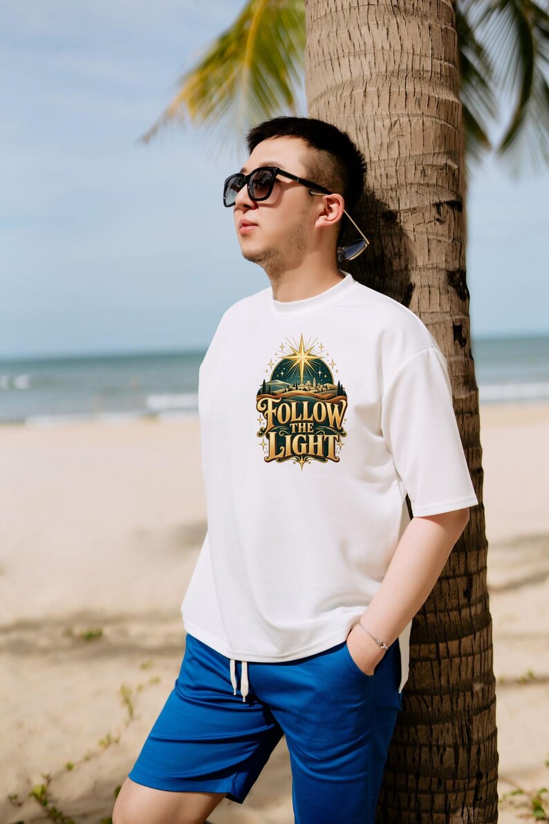 May include: A white short-sleeved t-shirt featuring a graphic print of a glowing star above a landscape with the text Follow The Light in gold and dark blue lettering. The shirt is paired with bright blue shorts and dark sunglasses.