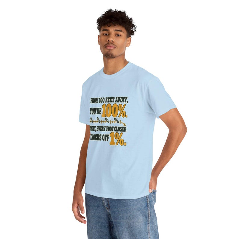 May include: A light blue short-sleeve t-shirt featuring the black and yellow text: FROM 100 FEET AWAY, YOU'RE 100%. SADLY, EVERY FOOT CLOSER KNOCKS OFF 1%. A graphic of a measuring line with footprints is positioned between the text lines.